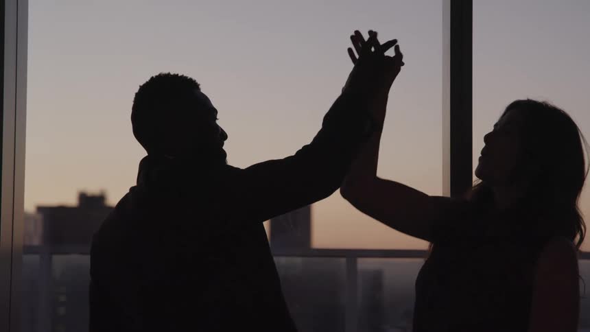 Two people in an office building share a high five in front of a large window overlooking a city scape