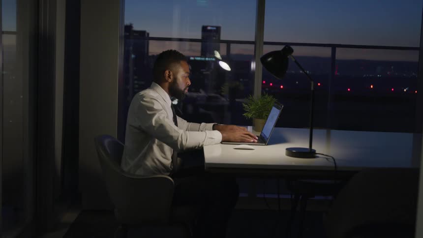 Male sit back in his chair at a table in an office at night fall leans forward to type on the laptop and then leans back again