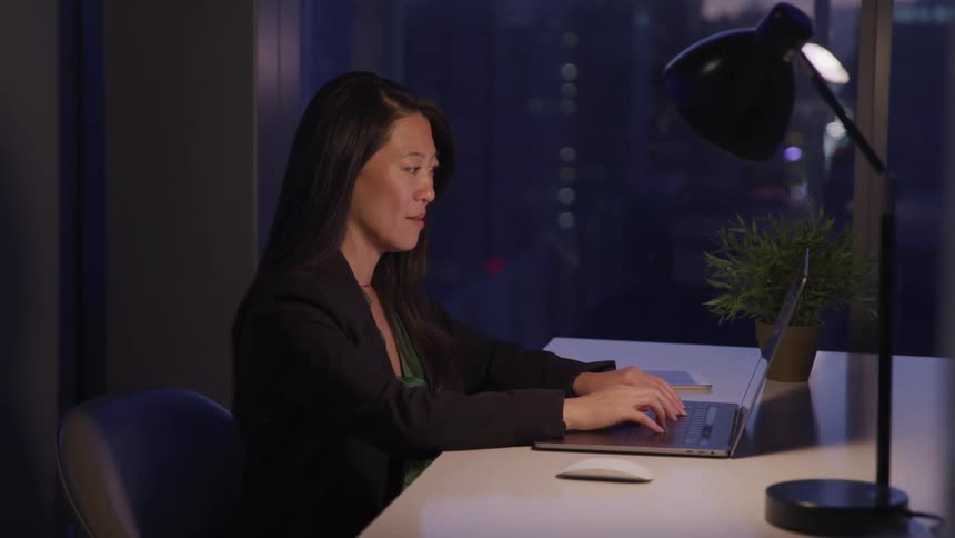 A middle aged asian woman sits in an office at night while typing on a laptop