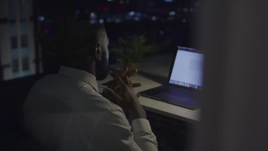 A man sits at a window view desk with a laptop at night