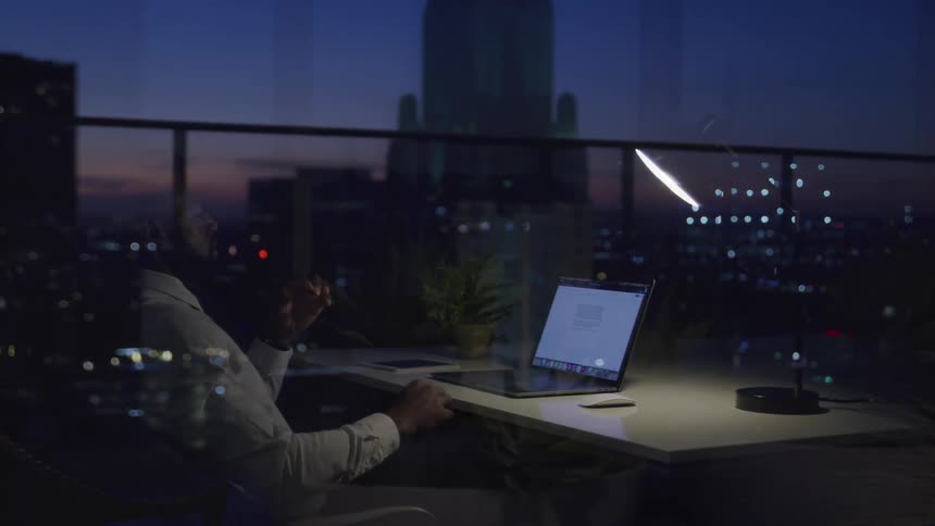 Black businessman with a beard sitting at a desk uses a laptop and works until late in the office at dusk