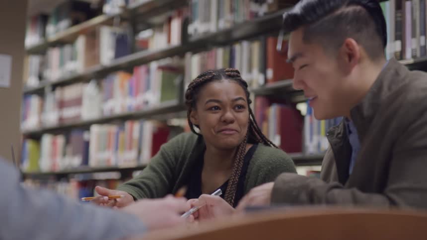 Three people sit at a table in a library and laugh as they talk together