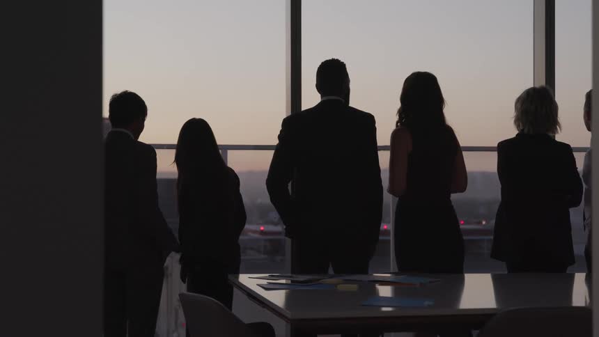 Business people looking out the windows are silhouetted against the sky at dusk in a conference room