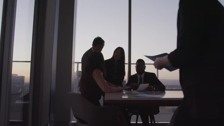 Two men and two women in a conference room around a table looking at a paper while a man and a woman walk around