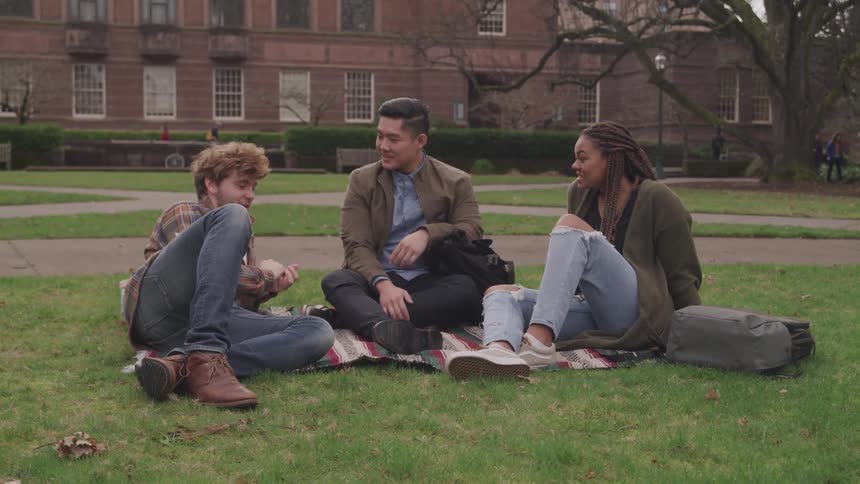 Two men and a woman sit on the yard outside of a college campus with their backpacks and books on the ground