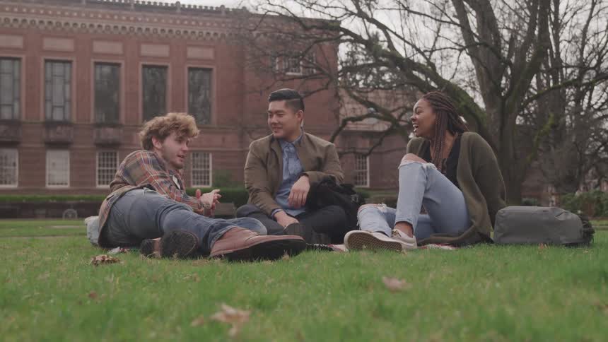 Asian man white man and black woman sit on green grass and talk to each other in the university yard