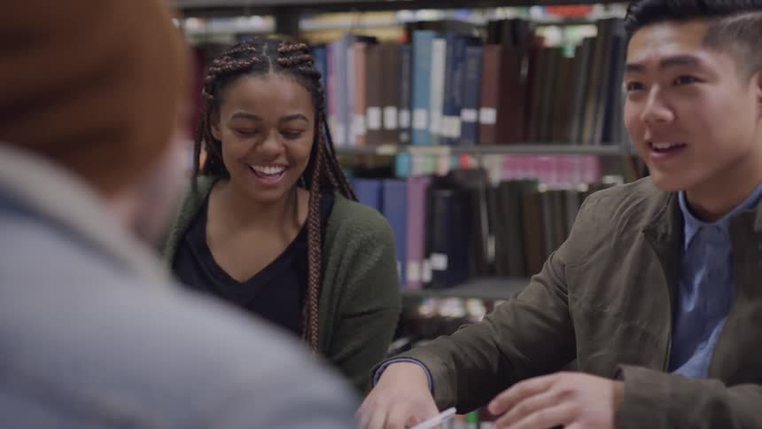 Black girl with braided hair asian man and white man sitting at a table talk to each other and laugh together in the public library