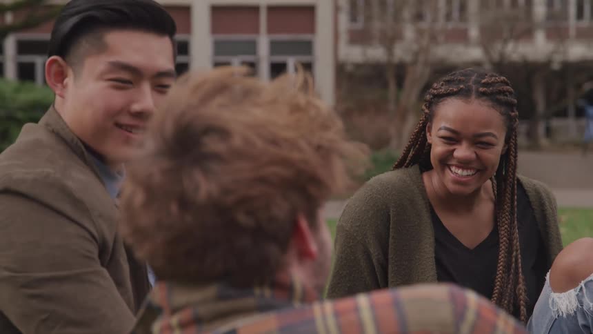 Asian man white man and black woman sit on green grass and laugh together in the university yard