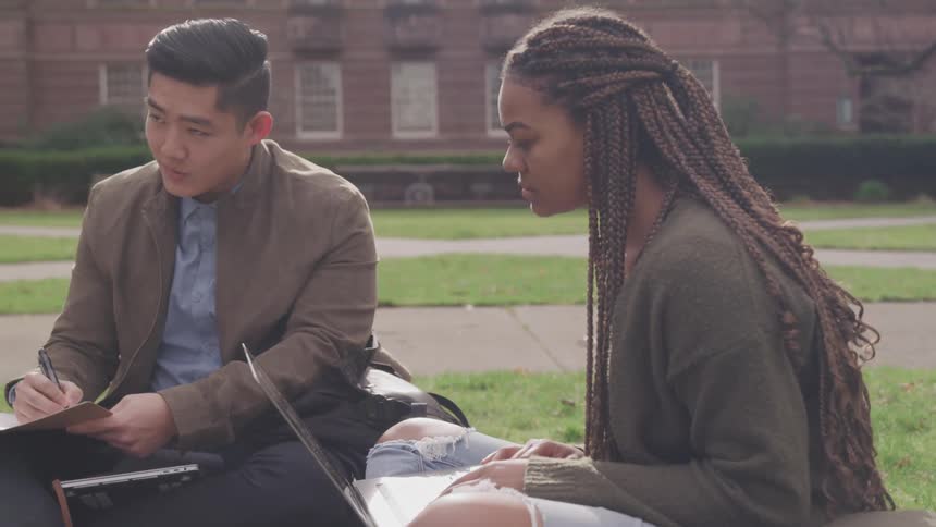 A woman sitting outside typing on a laptop next to a man with a pen in his hand writing