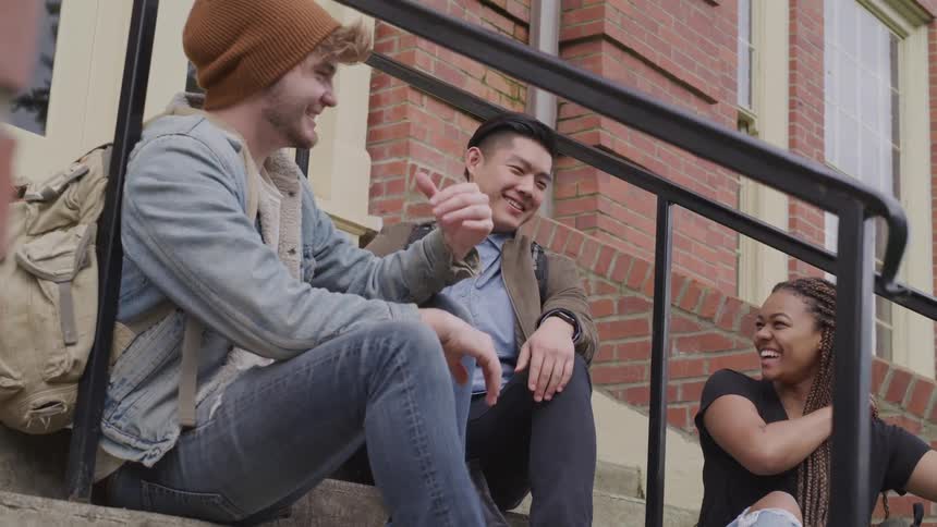 Three friends sit on the front steps of a brick building talking and laughing together