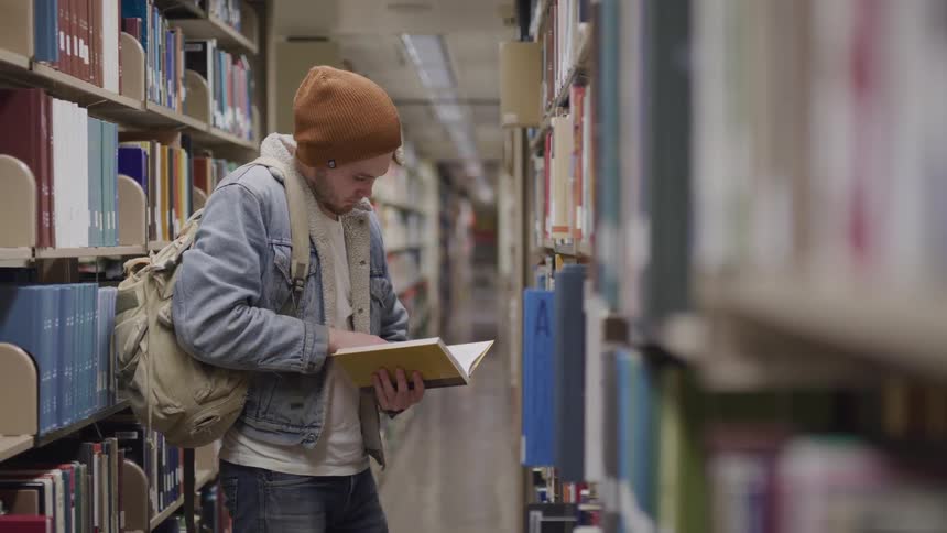 A wearing a backpack reads a book while standing in the aisle of a library