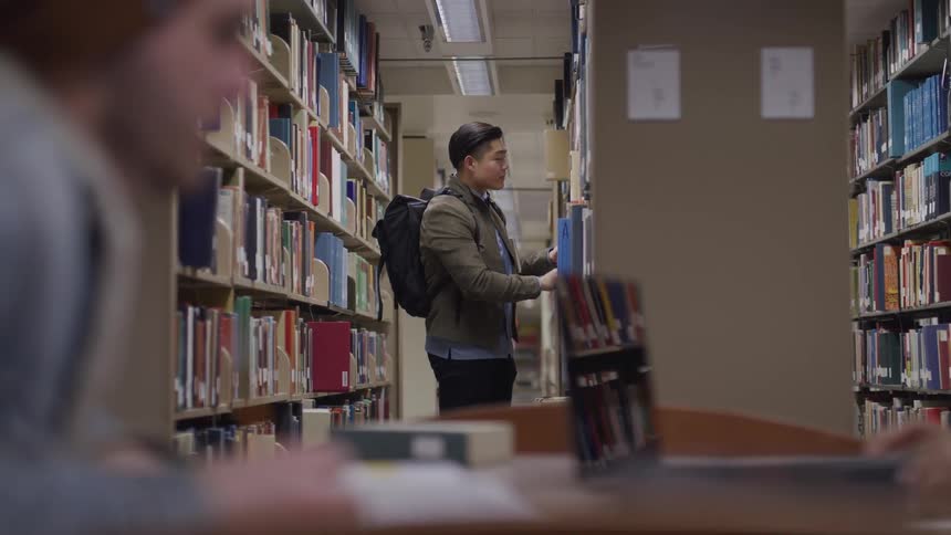 Asian man with a backpack and a green coat searches for a book in the library between bookshelves