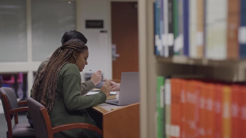 Three teenagers sit at a table in a school library and look at a laptop