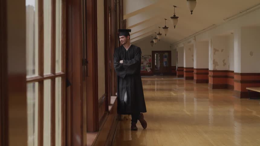 A man in a graduate gown and cap leans against a window frame in a school hallway