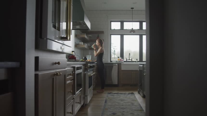 Woman with brown hair black sports sleeveless top and tights drinks a glass of smoothie in the kitchen
