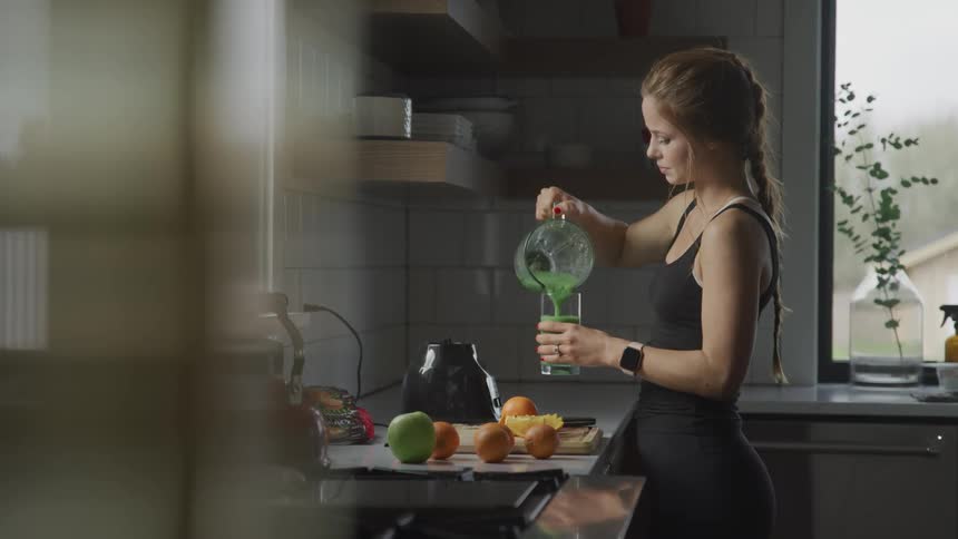 Woman with brown hair and a wristwatch pours a smoothie made from fruit and vegetables into a glass in the kitchen