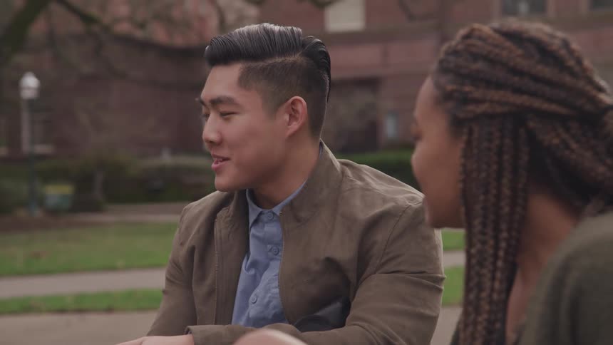 A black female and an asian male sitting down out a building chatting