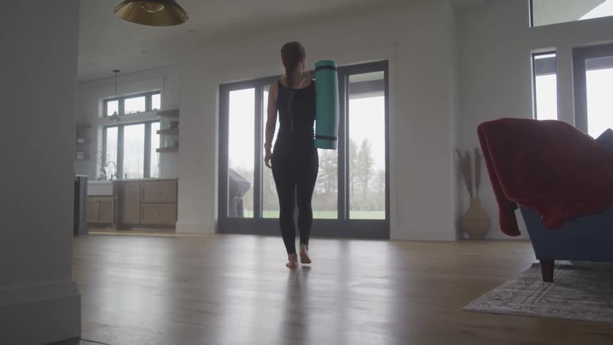 A woman carries a yoga mat through the living room of a house towards a sliding glass door