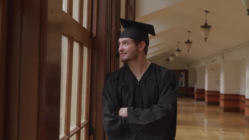 A young man smiles as he looks out a window with his cap and gown onfor graduation