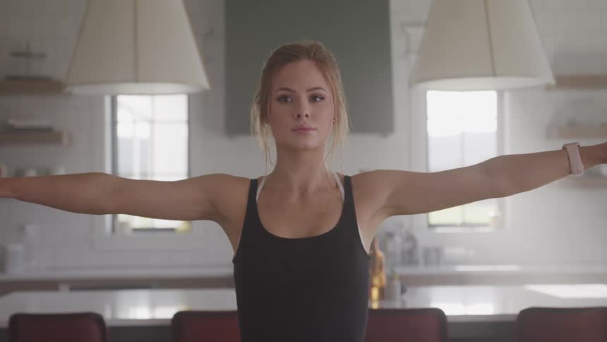 A young woman standing in a kitchen is performing a stretching exercise with her arms