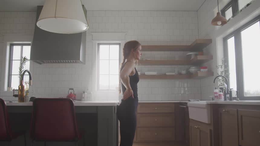 A woman in athletic wear takes deep breaths while standing against a counter in a kitchen