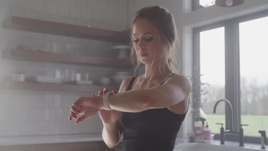 A woman in the kitchen looking at her wristwatch with a sink and a window behind her