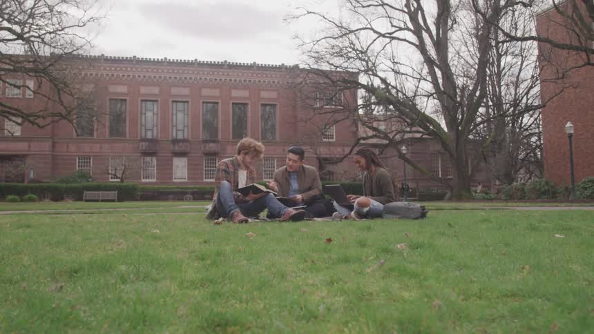 Three teens sit outside a school and read books and work on a laptop