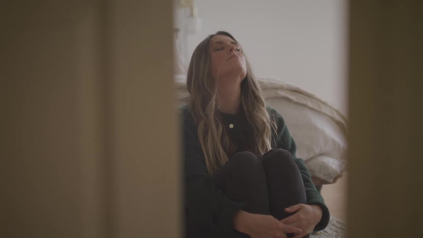A woman hugs her legs while sitting on the floor next to her bed