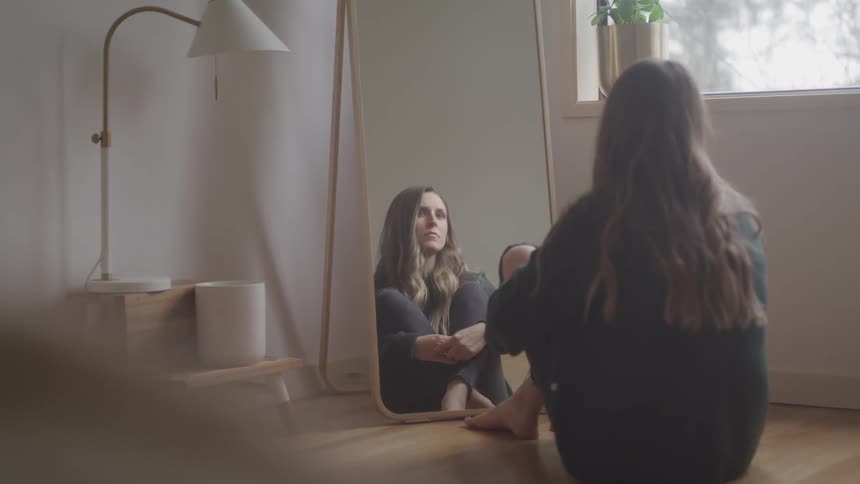 Young woman sitting on the floor of a room at home looks at her reflection in the mirror