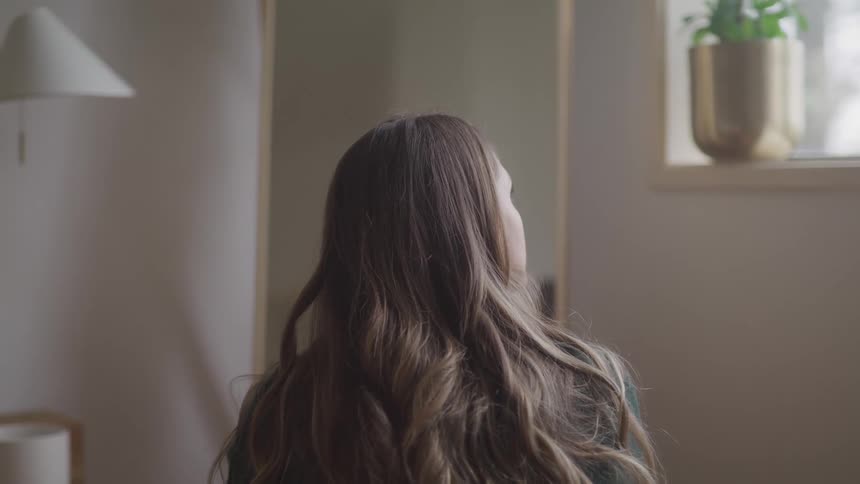 A woman sits on the floor of her bedroom in front of a mirror and looks out the window
