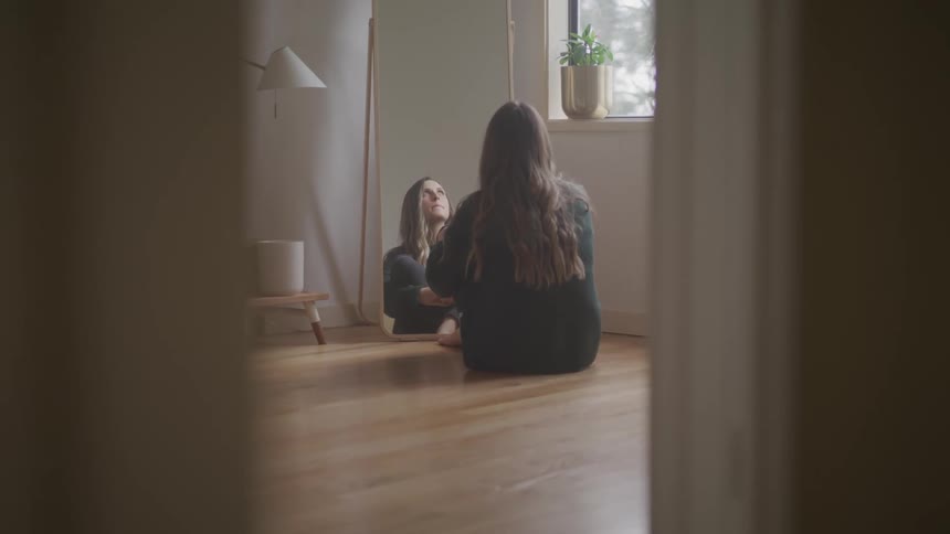 A woman sits alone in the corner of a room looking at a tall mirror