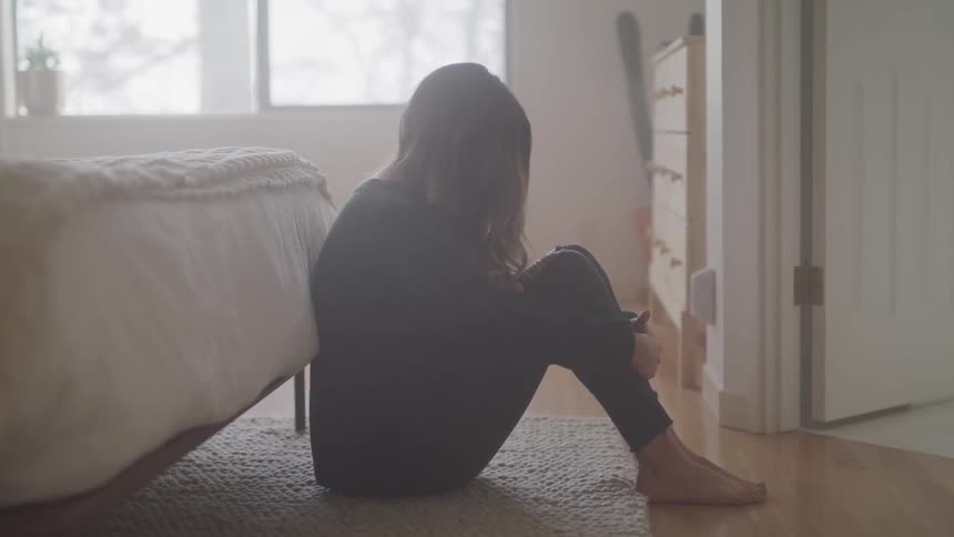 Woman with brown hair sits on the floor and leans against bed in the bedroom