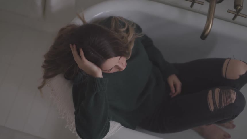 A sad woman sits fully clothed in a bathtub