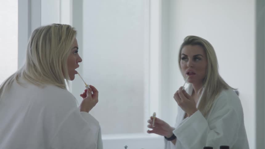A woman applies lipstick while standing in front of a large mirror in a bathrobe