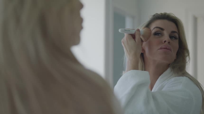 A woman in a bath robe looks at a mirror as she applies makeup to her face with a makeup brush