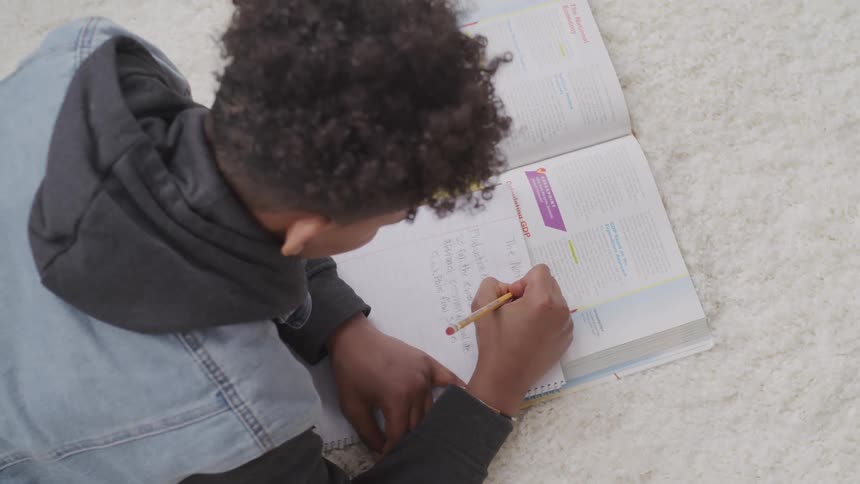 A young black man lies on a carpet as he reads a textbook and takes notes