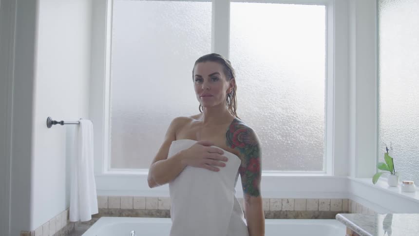 Tattooed woman with blondehair and a white towel faces the camera with eye contact in the bathroom
