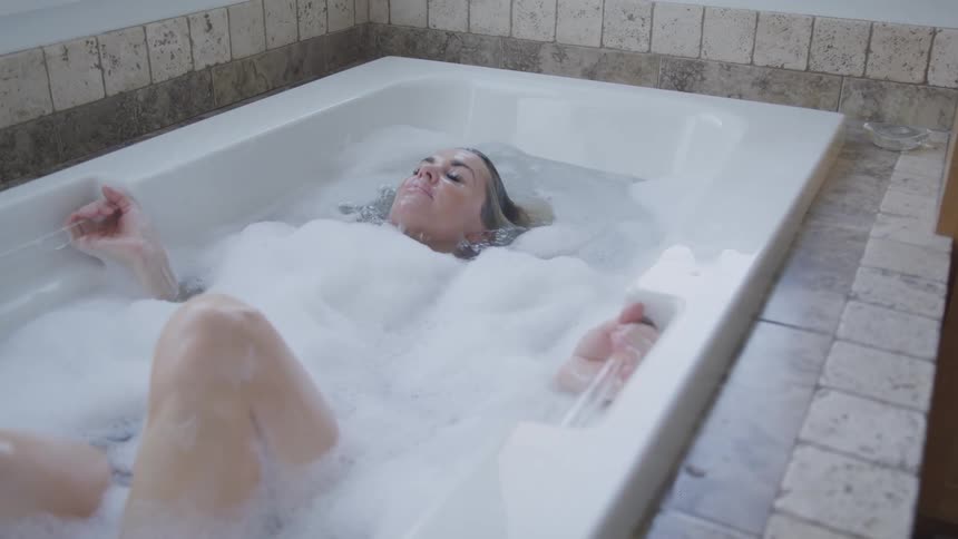 A tattooed woman lying under the water in a foamy bathtub slowly sits up