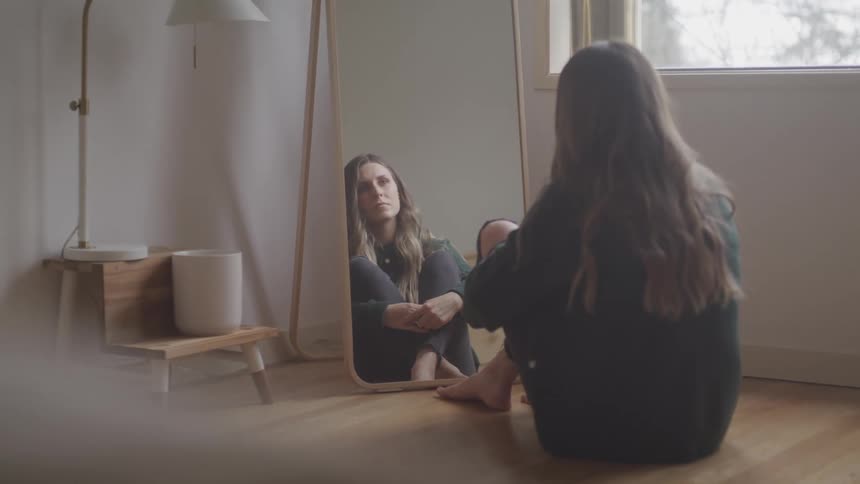 A woman sits alone in the corner of a room staring at a tall mirror