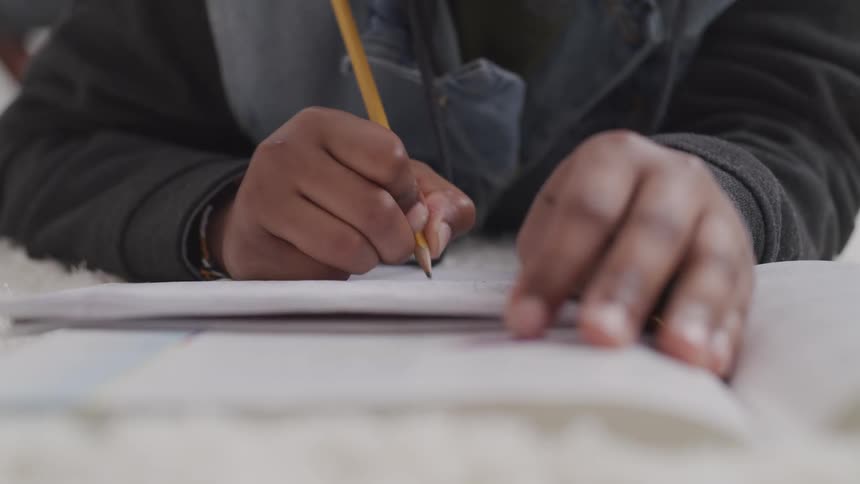 Black teenage boy sits at a table and writes on a notebook with a pencil