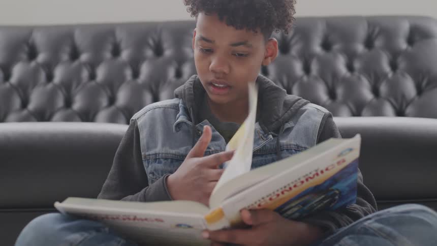 A boy sits on the floor in front of his couch and reads an economics textbook