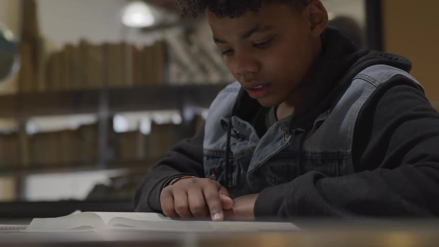 Black teenage boy sits at a table and reads a book in the public library