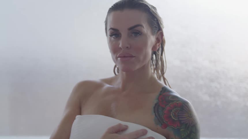 Middle aged blonde woman with a tattoo on her shoulder stands wrapped in towel in the bathroom