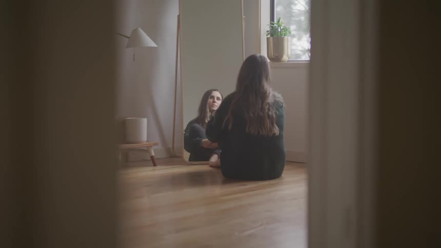 Woman with a green pullover sits on the floor and looks at a tall dressing mirror