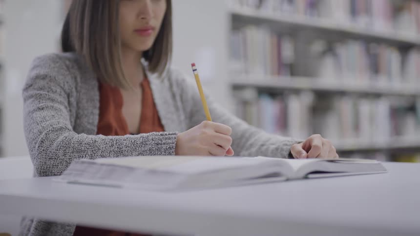 A young asian woman writes in her notebook while sitting with a large book at a table in the library