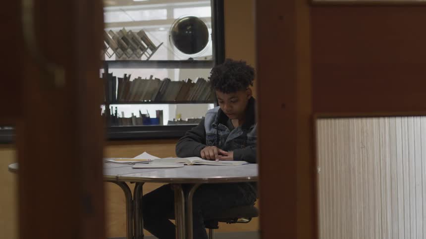 A boy traces the words of a textbook with his finger and he studies at a table