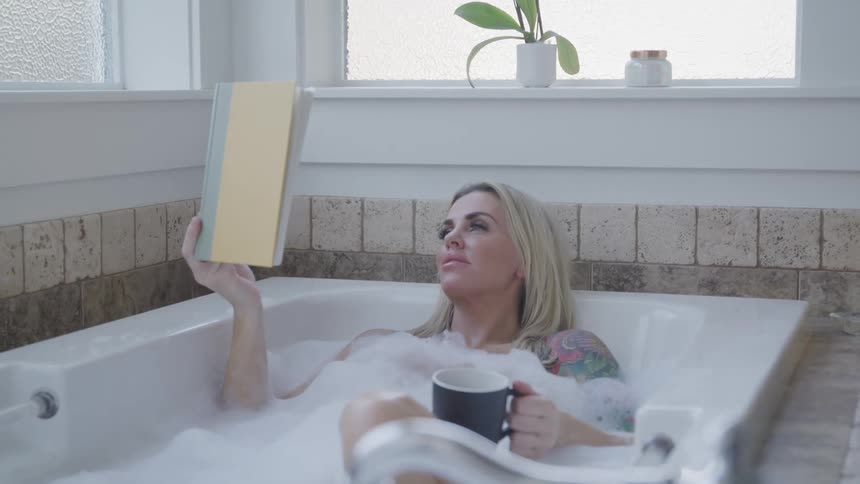 A woman in a bathtub with bubbles holding a mug and reading a book