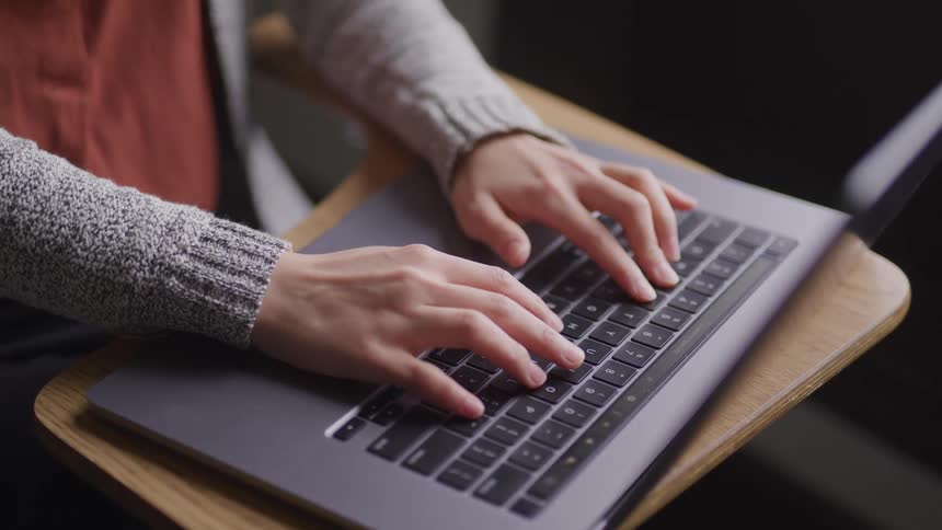 A person types on a laptop while sitting in a classroom desk