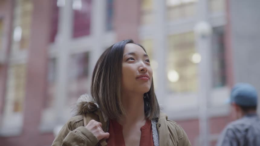 Teenage asian girl with brown coat smiles slightly and looks up at buildings in the street