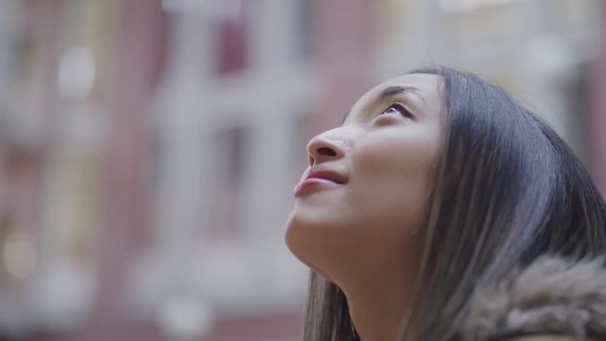 Teenage asian girl with straight hair and a brown coat looks up at buildings in the street